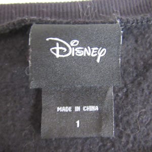 Peut inclure: Gros plan sur une &eacute;tiquette de v&ecirc;tement noire avec le logo Disney en blanc. En dessous, une petite &eacute;tiquette indique "MADE IN CHINA" et le chiffre "1". Le tissu est une mati&egrave;re sombre et textur&eacute;e.