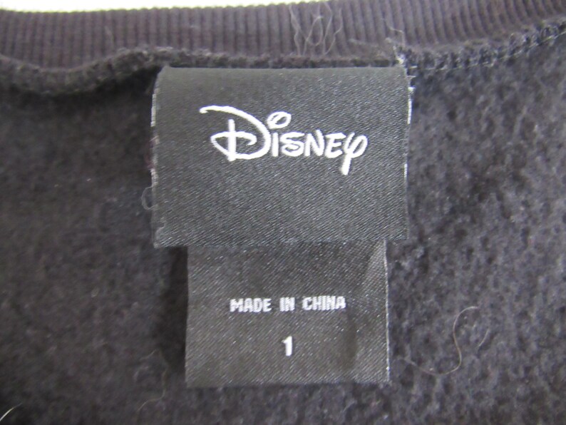 Peut inclure: Gros plan sur une &eacute;tiquette de v&ecirc;tement noire avec le logo Disney en blanc. En dessous, une petite &eacute;tiquette indique "MADE IN CHINA" et le chiffre "1". Le tissu est une mati&egrave;re sombre et textur&eacute;e.