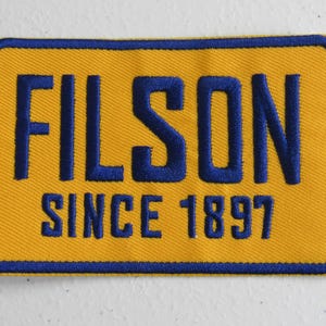 Könnte beinhalten: Ein gelber Stoff-Patch mit einem blauen gestickten Rand. Der Patch zeigt das Wort "FILSON" in großen blauen Buchstaben und den Text "SINCE 1897" in kleineren blauen Buchstaben.