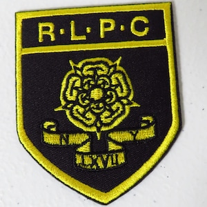 Puede incluir: Parche bordado en negro y amarillo con las letras "R.L.P.C" en la parte superior. El parche presenta un emblema floral y el número romano "LXXVII" en la parte inferior. El parche tiene un borde amarillo.