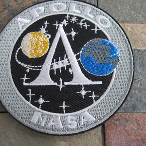 Może przedstawiać: Naszywka haftowana na biało z srebrnym tłem. Naszywka przedstawia logo NASA z napisami "NASA" na dole i "APOLLO" na górze. Logo zawiera również białą literę "A" z żółtym księżycem i niebieską Ziemią po obu stronach litery. Naszywka jest otoczona srebrną obwódką.