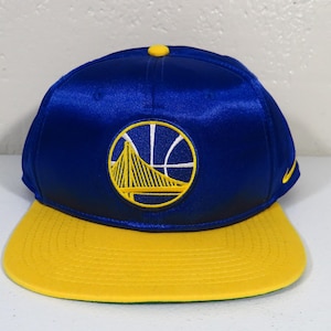 Puede incluir: Una gorra de béisbol azul y amarilla con el logotipo de los Golden State Warriors. La gorra tiene una visera amarilla y una corona azul con el logotipo del equipo bordado en la parte delantera. Un pequeño botón amarillo está en la parte superior.