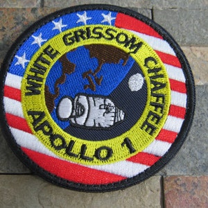 Puede incluir: Un parche redondo bordado con un diseño amarillo, negro y azul. El parche presenta una cápsula blanca con una bandera estadounidense roja, blanca y azul en el fondo. El texto "WHITE GRISSOM CHAFFEE APOLLO 11" está bordado en amarillo alrededor de la cápsula.