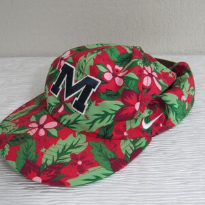 May include: A red, green, and white floral print baseball cap with a large navy blue and white letter "M" on the front. The Nike swoosh logo is visible on the side.