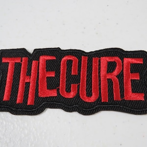 May include: A rectangular embroidered patch with a black background and the text THE CURE stitched in bold red thread. The patch features a thick black border that follows the irregular, slightly jagged contour of the lettering.