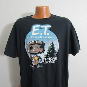 Pode incluir: Camiseta preta com uma imagem de desenho animado de E.T. o extraterrestre segurando um dedo brilhante. A imagem está sobre um fundo de lua azul com um pinheiro no fundo. O texto "Phone Home" está impresso abaixo da imagem.
