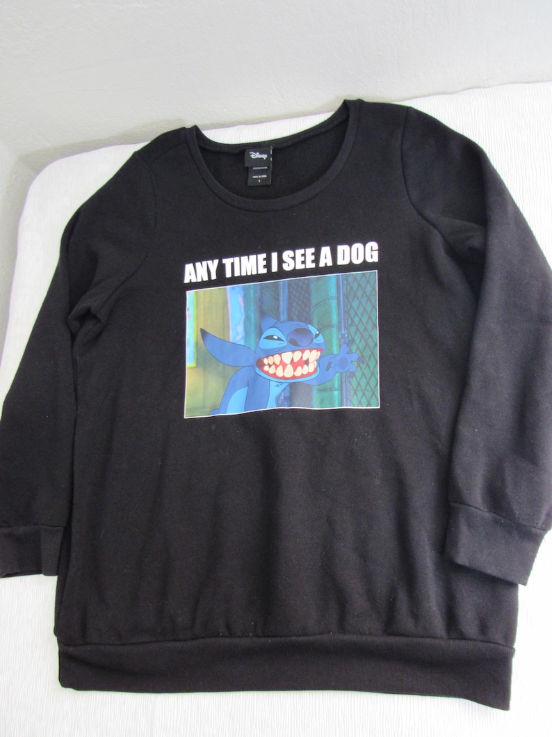 Peut inclure: Sweat-shirt noir &agrave; manches longues avec un dessin du personnage Disney Stitch et le texte "ANY TIME I SEE A DOG". L'image montre Stitch souriant. Le sweat-shirt a un col rond.