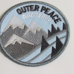 Può includere: Una toppa ricamata blu e grigia con il testo "Outer Peace" e "Arc'teryx" in bianco.
