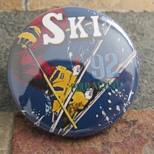 May include: A blue and white button with a skier in red, yellow, and blue clothing. The button says "SKI-92" in white letters.