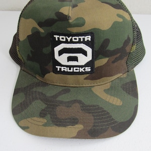 May include: Camouflage trucker hat with a black and white patch that says "TOYOTA TRUCKS" in white letters.