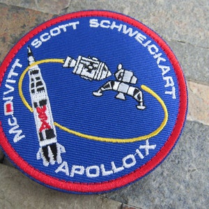 May include: A blue and red embroidered patch with a white rocket and a white lunar module orbiting each other. The text "McDivitt, Scott, Schweickart, Apollo IX" is around the edge of the patch.