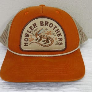 May include: Orange trucker hat with a tan mesh back and a curved brim. The front features a patch with the words "HOWLER BROTHERS" around a stylized lizard illustration. A white rope detail accents the brim.