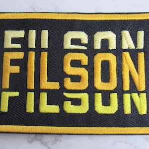May include: A black patch with a yellow border and the word "FILSON" embroidered in yellow letters.