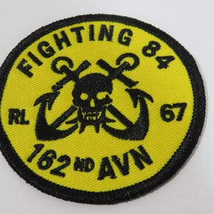 May include: A circular embroidered patch with a bright yellow background and a black border. The design features a black skull centered over two crossed anchors. The text reads FIGHTING 84, RL, 67, and 162 ND AVN in black embroidery.