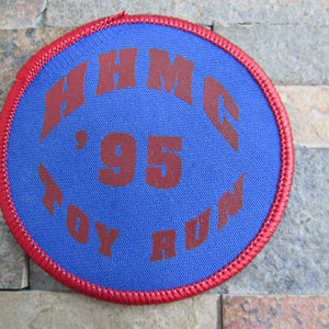 Puede incluir: Un parche circular azul y rojo con el texto "BHMC '95 TOY RUN" en marrón.