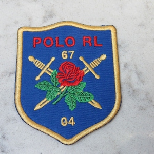 Op de afbeelding: Geborduurde blauwe patch met gouden rand, met de tekst "POLO RL 67 04". Het ontwerp bevat gekruiste zwaarden, een rode roos en groene bladeren. De patch heeft een schildvorm.