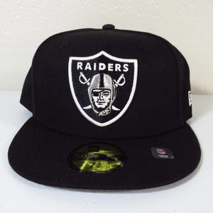 Puede incluir: Gorra de béisbol negra con el logotipo de los Raiders en blanco. El logotipo es un escudo con el nombre del equipo y una cara con casco. La gorra tiene una visera plana y un diseño ajustado, para los fanáticos del deporte.