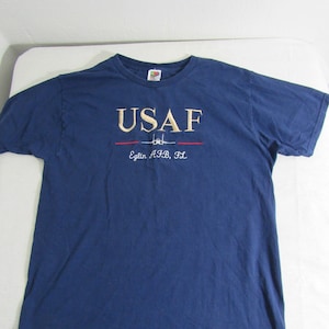 Op de afbeelding: Een marineblauw t-shirt met een goud geborduurd "USAF" logo en de tekst "Eglin AFB, FL" in rood.