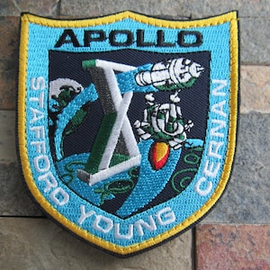 Puede incluir: Un parche bordado azul y blanco con el texto "APOLLO" en la parte superior, "STAFFORD YOUNG CERNAN" alrededor de los bordes y una representación de un cohete lanzándose al espacio.
