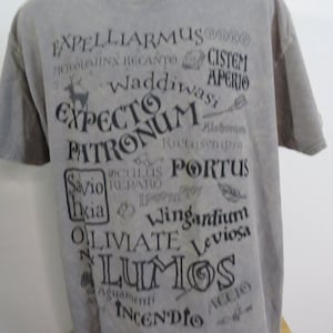 Puede incluir: Camiseta gris con texto negro con varios hechizos y frases. La camiseta incluye las palabras "Expecto Patronum", "Lumos" y "Expelliarmus", entre otras. También es visible un gráfico de ciervo.