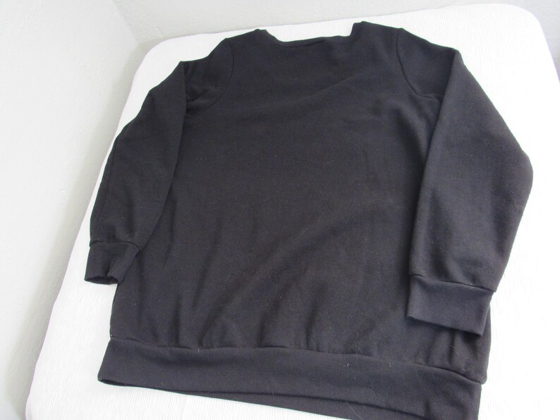 Peut inclure: Un sweat-shirt noir &agrave; manches longues avec un col ras du cou et des poignets et un ourlet c&ocirc;tel&eacute;s. Le sweat-shirt est fait d'un tissu uni et doux. Le v&ecirc;tement est pos&eacute; &agrave; plat sur une surface blanche.