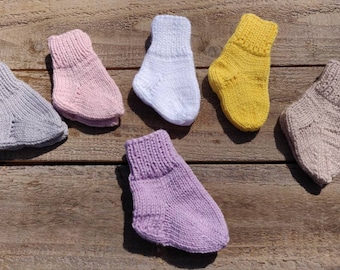 May include: Six pairs of knitted baby socks in different colors: gray, pink, white, yellow, purple, and beige. The socks are laid out on a wooden surface.