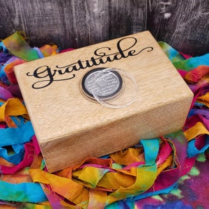 May include: A wooden box with the word "Gratitude" in a cursive font on the lid. A silver coin with text is on top, covered by a clear plastic circle. The box sits on a colorful fabric with various hues.