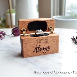 K&ouml;nnte beinhalten: Eine rechteckige Ringbox aus Mahagoniholz mit Klappdeckel, auf deren Vorderseite der eingravierte Text "I'LL LOVE YOU Always" zu sehen ist. Das Innere ist mit schwarzem Samt ausgekleidet und enth&auml;lt einen silberfarbenen Ring mit Stein.