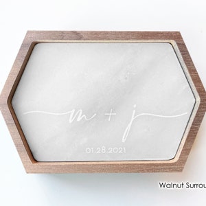 May include: A hexagonal ring box with a walnut surround and a gray concrete insert. The insert is engraved with the text "m + j" and the date "01.28.2021".