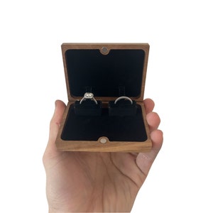 May include: A wooden ring box with a black velvet interior. The box is open and two rings are visible inside.