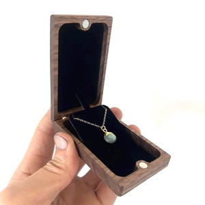 May include: A dark brown wooden jewelry box with a hinged lid and magnetic closure, featuring a black velvet interior lining. Inside the box rests a delicate gold chain necklace with a teardrop-shaped, grey-green gemstone pendant with a gold cap.