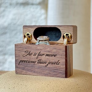 May include: A rectangular wooden ring box with a hinged lid featuring gold-tone metal hinges. The front of the box is engraved with the text "She is far more precious than jewels." Inside, a silver-toned ring with a central stone is displayed.