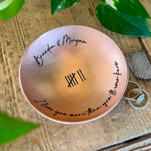 May include: A round copper-colored trinket dish featuring black engraved text that reads Brandon & Morgan, a tally mark symbol, and the phrase I love you more than you'll ever know. Two silver-toned rings rest on the edge of the dish.