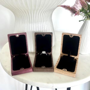 May include: Three rectangular wooden ring boxes with hinged lids and magnetic closures. Each box features a black velvet interior lining. The center box contains a silver ring with a central stone. The boxes are purple, dark brown, and light tan.