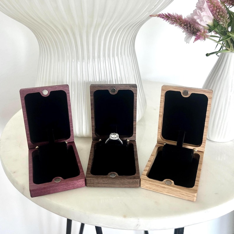 May include: Three rectangular wooden ring boxes with hinged lids and magnetic closures. Each box features a black velvet interior lining. The center box contains a silver ring with a central stone. The boxes are purple, dark brown, and light tan.