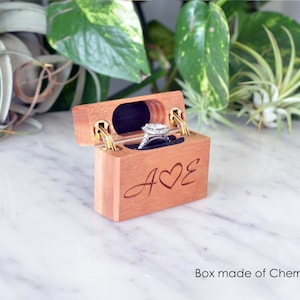 K&ouml;nnte beinhalten: Eine rechteckige Ringbox aus Kirschholz mit Klappdeckel und goldfarbenen Metallscharnieren. Die Vorderseite ist mit den Buchstaben A und E graviert, die durch ein Herz getrennt sind. Innen h&auml;lt ein dunkelblauer Samteinsatz einen Silberring.