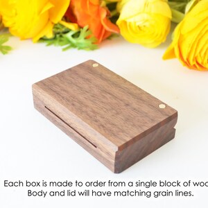 May include: A rectangular dark brown wooden box with a lid featuring two small circular brass inlays. The text below reads: Each box is made to order from a single block of wood. Body and lid will have matching grain lines.
