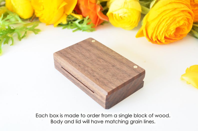 May include: A rectangular dark brown wooden box with a lid featuring two small circular brass inlays. The text below reads: Each box is made to order from a single block of wood. Body and lid will have matching grain lines.
