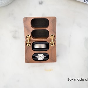 May include: A wooden ring box with two compartments, one for an engagement ring and one for a wedding band. The box is made of walnut wood and has gold hinges. The ring box is open and the rings are visible.