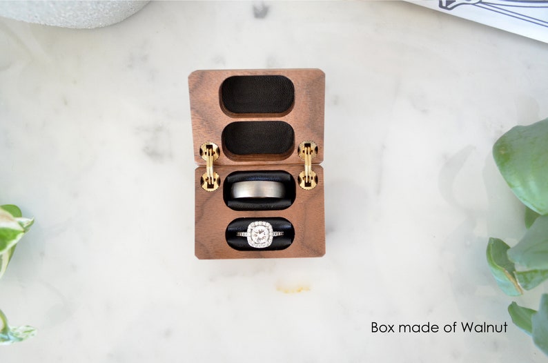 May include: A wooden ring box with two compartments, one for an engagement ring and one for a wedding band. The box is made of walnut wood and has gold hinges. The ring box is open and the rings are visible.