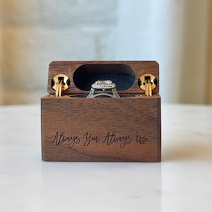K&ouml;nnte beinhalten: Eine rechteckige, dunkelbraune Holzschatulle f&uuml;r Ringe mit Klappdeckel und der eingravierten Aufschrift Always You, Always Us auf der Vorderseite. Die ge&ouml;ffnete Schatulle zeigt einen silberfarbenen Ring mit einem zentralen Stein im Inneren.