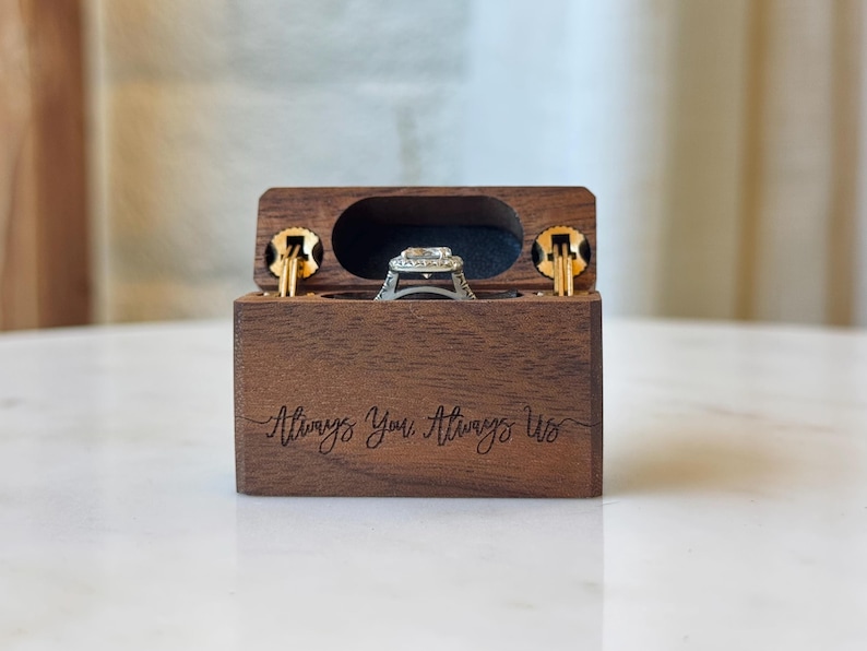 K&ouml;nnte beinhalten: Eine rechteckige, dunkelbraune Holzschatulle f&uuml;r Ringe mit Klappdeckel und der eingravierten Aufschrift Always You, Always Us auf der Vorderseite. Die ge&ouml;ffnete Schatulle zeigt einen silberfarbenen Ring mit einem zentralen Stein im Inneren.
