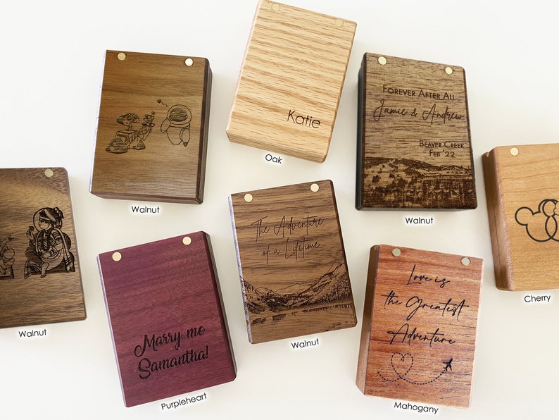 May include: Assortment of small wooden boxes with different wood types and engraved designs. The boxes feature various engravings, including a couple, a mountain scene, a heart with an airplane, and a Mickey Mouse silhouette. The wood types include walnut, oak, cherry, mahogany, and purpleheart.