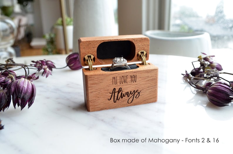 K&ouml;nnte beinhalten: Eine rechteckige Ringbox aus Mahagoniholz mit Klappdeckel, auf deren Vorderseite der eingravierte Text "I'LL LOVE YOU Always" zu sehen ist. Das Innere ist mit schwarzem Samt ausgekleidet und enth&auml;lt einen silberfarbenen Ring mit Stein.