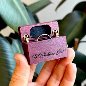 K&ouml;nnte beinhalten: Eine kleine rechteckige Ringbox aus violettem Holz mit goldfarbenen Metallscharnieren und schwarzem Innenfutter. Auf der Vorderseite der Box befindet sich die Gravur To Whatever End, und im Inneren des offenen Beh&auml;lters ist ein d&uuml;nner Goldring sichtbar.