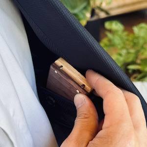 A hand slipping a compact wooden engagement ring box into the inside pocket of a suit jacket, demonstrating its ultra-slim, hidden profile.