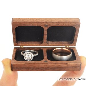May include: A rectangular walnut wood ring box with a hinged lid and two interior compartments lined with black fabric. The left compartment holds a silver-toned ring with a central stone and halo setting, while the right holds a plain metal band.