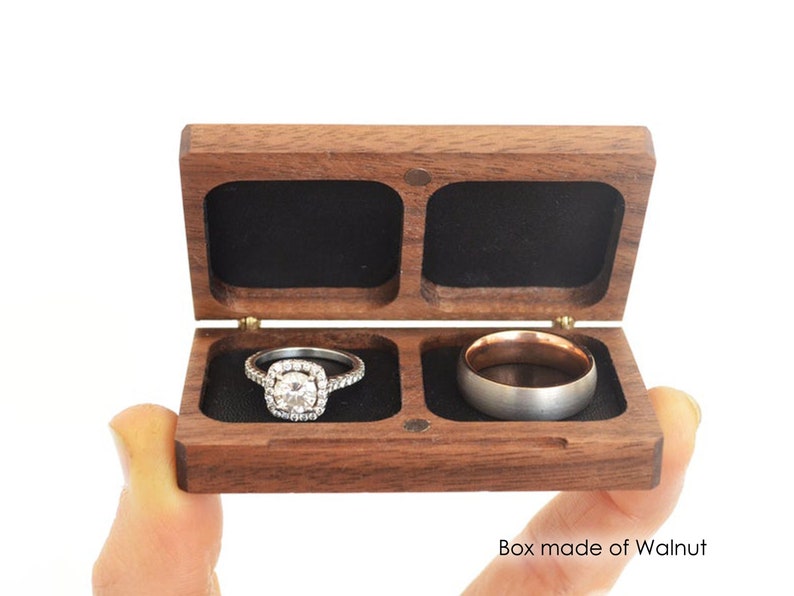 May include: A rectangular walnut wood ring box with a hinged lid and two interior compartments lined with black fabric. The left compartment holds a silver-toned ring with a central stone and halo setting, while the right holds a plain metal band.