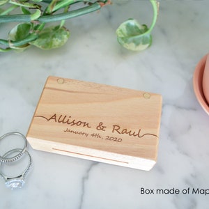 May include: A rectangular light-coloured wooden box with two circular magnetic closures. The lid is engraved with the text "Allison & Raul" and "January 4th, 2020" in a cursive script. Two silver-toned metal rings are positioned next to the box.