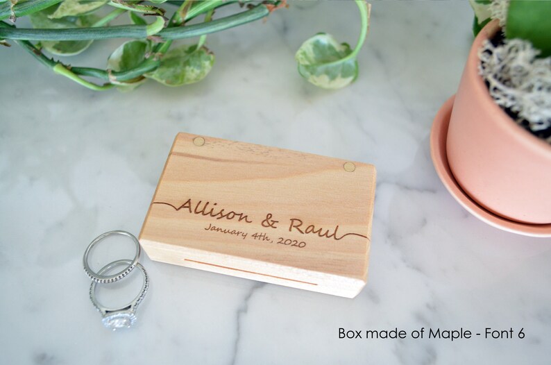 May include: A rectangular light-coloured wooden box with two circular magnetic closures. The lid is engraved with the text "Allison & Raul" and "January 4th, 2020" in a cursive script. Two silver-toned metal rings are positioned next to the box.